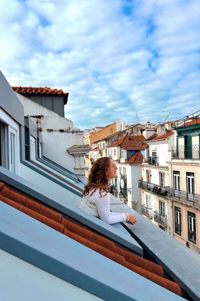 Traveler Liz leaning out from a slanted rooftop window, smiling while overlooking the colorful buildings and tiled roofs of Lisbon under a bright, cloudy sky.
