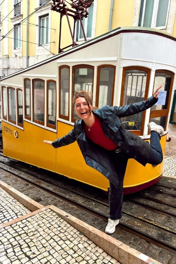 Traveler Liz playfully posing with one leg lifted beside Lisbon’s iconic yellow tram on a cobblestone street.