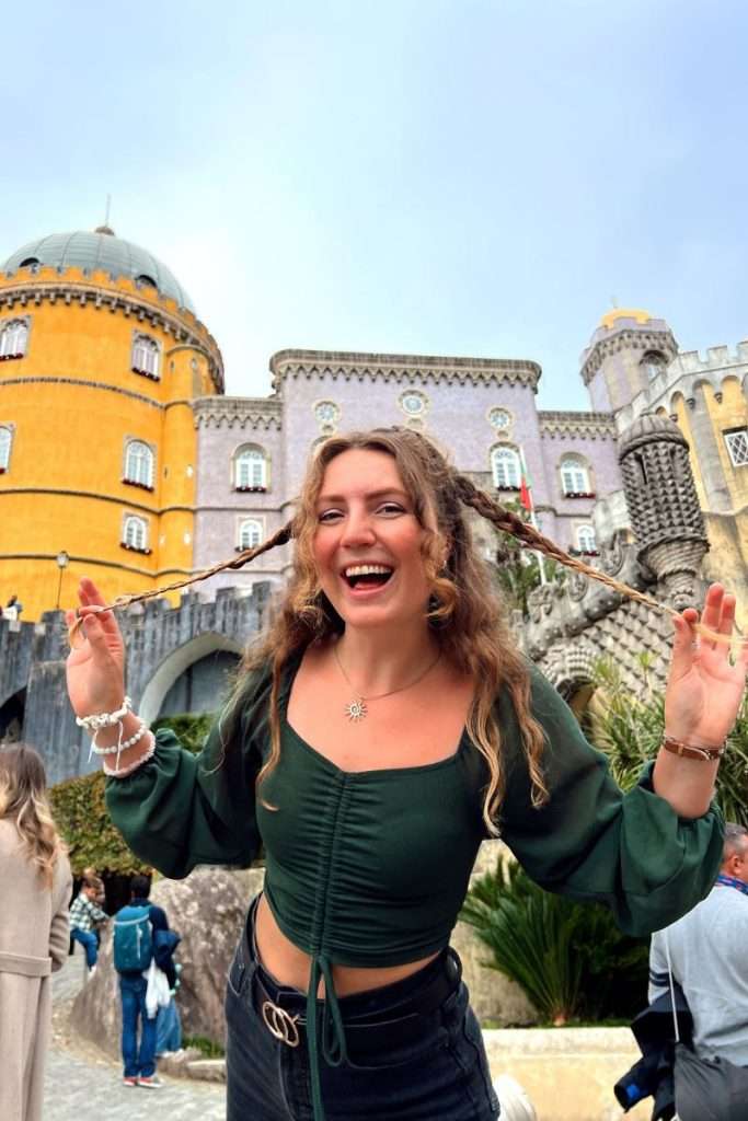 Traveler Liz smiling and playfully holding her braided hair in front of the colorful exterior of Pena Palace in Sintra, Portugal.