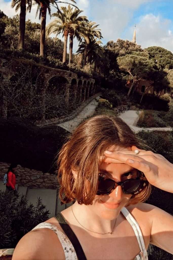 Traveler Liz taking a selfie in the sun at Park Güell in Barcelona, with palm trees and Gaudí’s stone arches in the background.