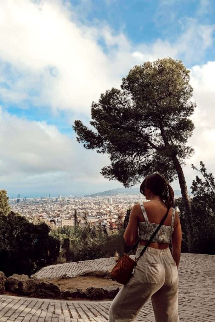 Traveler Liz looking out over Barcelona from a viewpoint in Park Güell, surrounded by trees and panoramic city views.