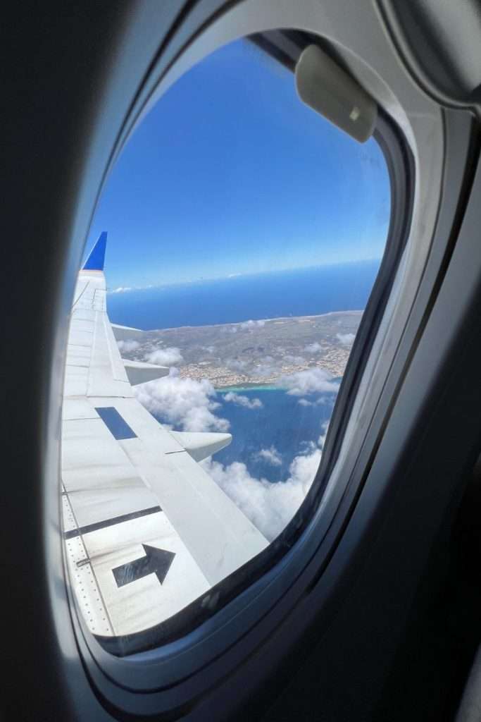 View from an airplane window while flying over Aruba, showing the wing, turquoise coastline, and blue sky above the island.