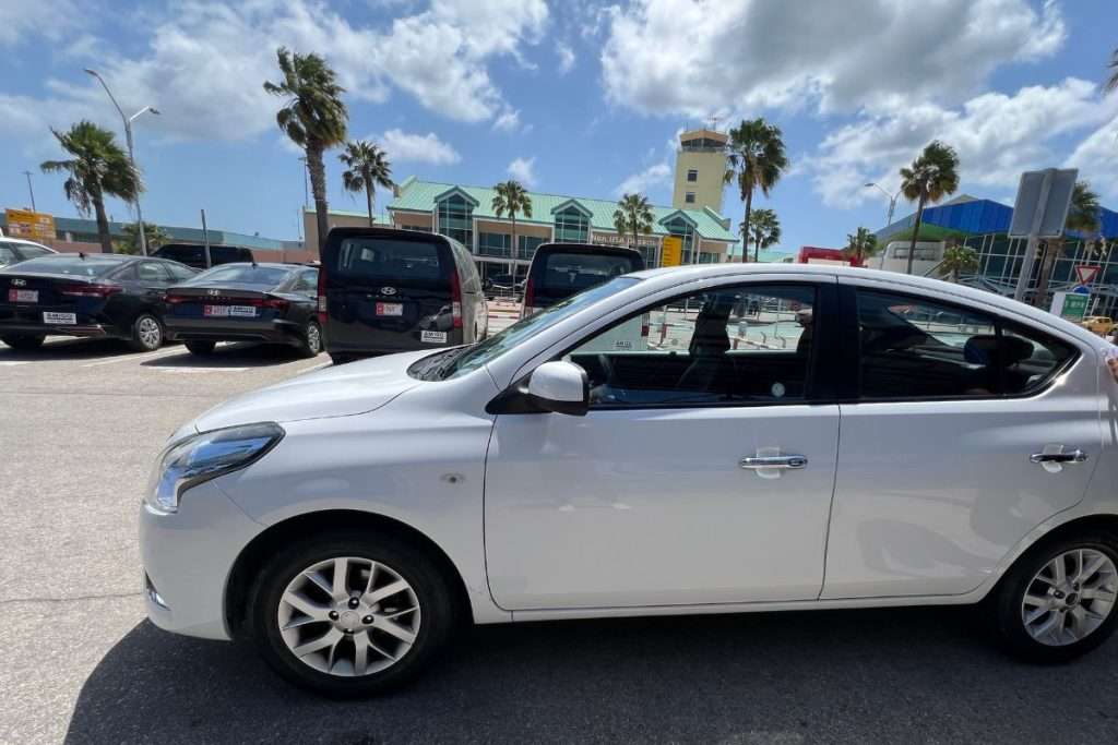 Rental car parked outside the Aruba airport, with palm trees and the terminal building visible in the background on a sunny day.