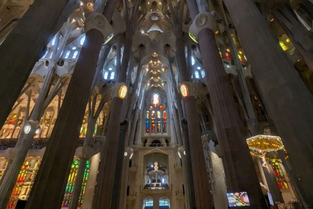Interior view of La Sagrada Familia in Barcelona, showing tall columns, intricate ceiling designs, and colorful stained glass windows.