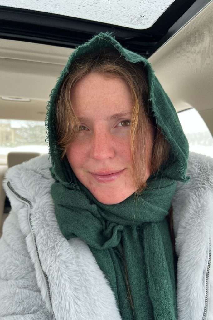 Liz sitting in a car wearing a warm winter coat and green scarf wrapped over her head, bundled up for cold Colorado weather during a ski trip.