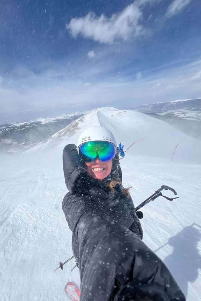 Woman taking a selfie while skiing at the top of a snowy peak in Breckenridge, Colorado, wearing a white helmet and reflective goggles on a windy winter day.