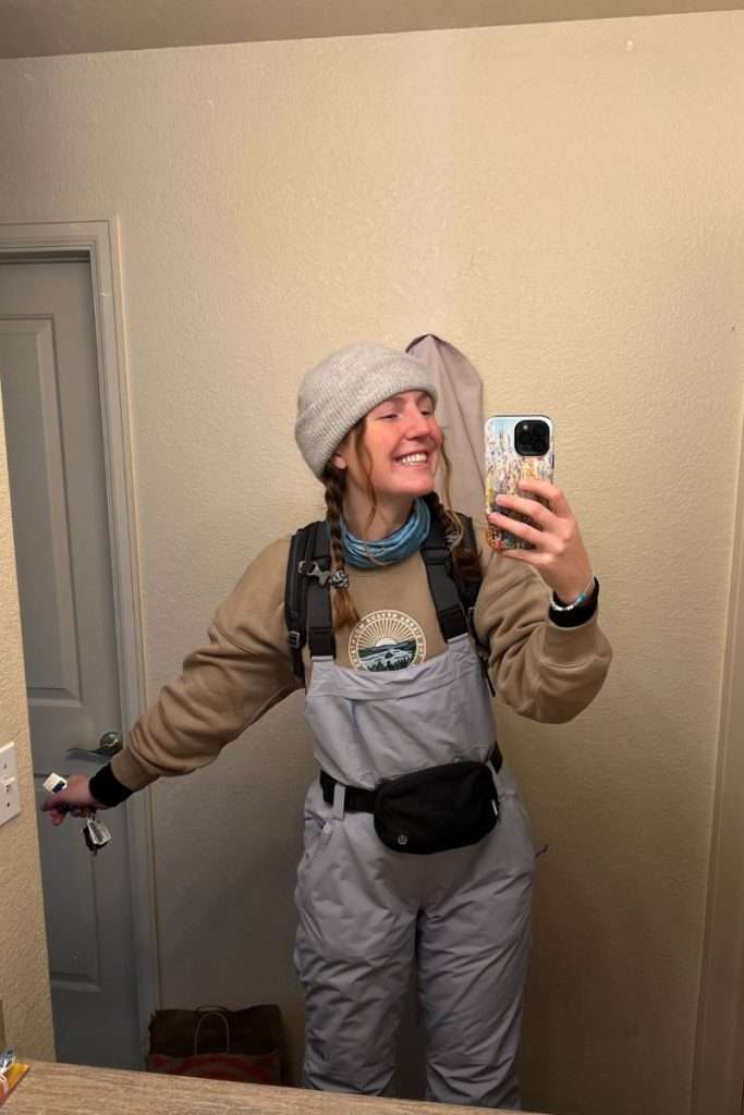 Woman smiling in a mirror wearing ski bibs, a sweatshirt, beanie hat, and fanny pack: an example of layering for a Colorado ski trip packing list.