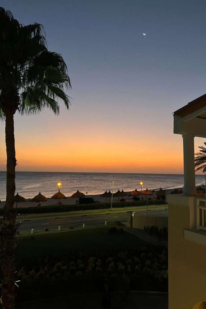 Sunset over the beach in Aruba with palapas, palm trees, and the ocean glowing orange beneath a crescent moon, viewed from a balcony.