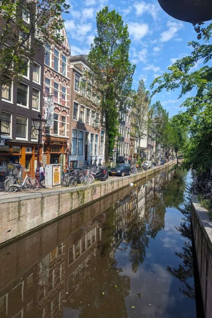 Amsterdam canal lined with historic narrow buildings, parked bicycles, and leafy trees, with calm water reflecting the houses on a bright, sunny day.