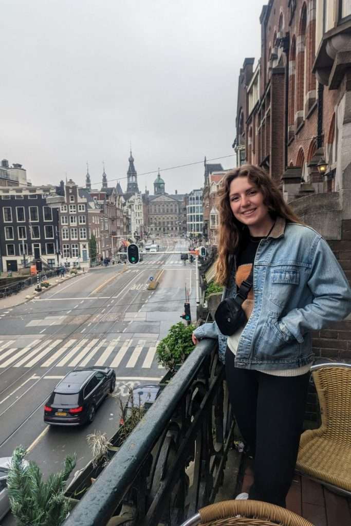 Smiling woman standing on a small balcony overlooking an Amsterdam street, with historic buildings, tram tracks, and a city square visible in the background on a cloudy day.