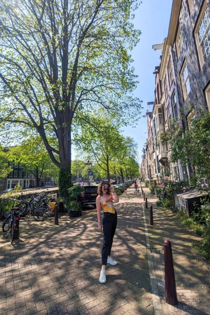 Woman walking along a quiet Amsterdam street lined with tall canal houses, leafy trees, parked bicycles, and brick sidewalks on a sunny spring day.