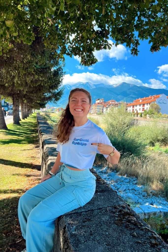 Traveler Liz wearing an Our Great Perhaps shirt sits on a stone wall near a river, smiling with mountains and buildings in the background.