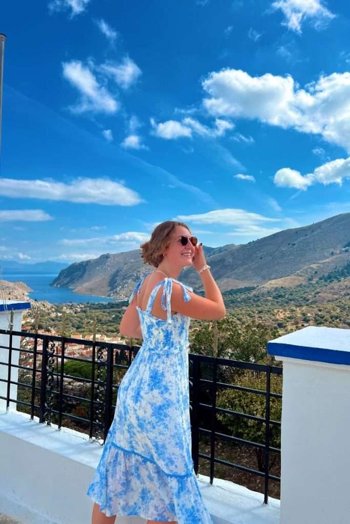A woman wearing a blue summer dress looks out over a scenic coastal landscape with mountains, blue water, and a bright sky while traveling.