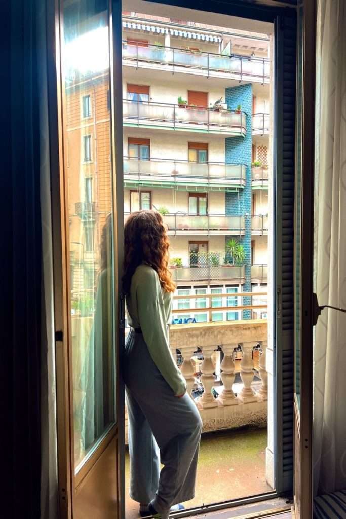 A woman stands in a doorway overlooking a city balcony and residential buildings, reflecting a calm and intentional solo travel moment.