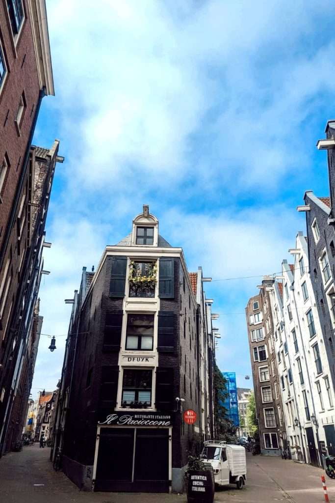 Narrow historic building on a quiet Amsterdam street with classic canal houses on both sides, blue sky overhead, and an Italian restaurant at street level.