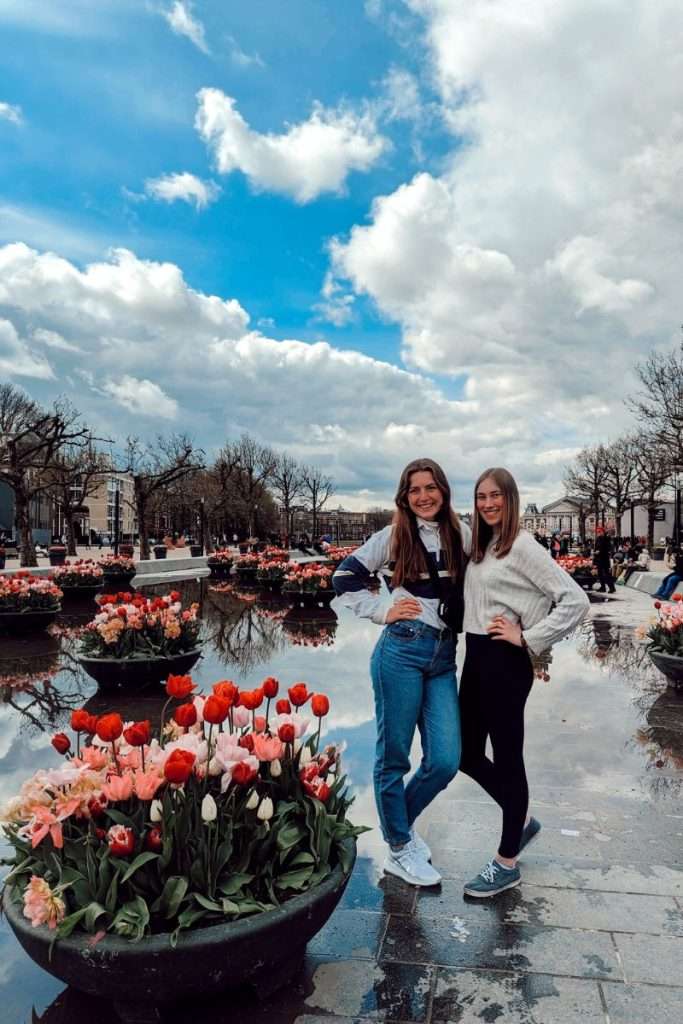 Liz and Amy near the museums in Amsterdam surrounded by tulips with a wet ground reflecting the sky.