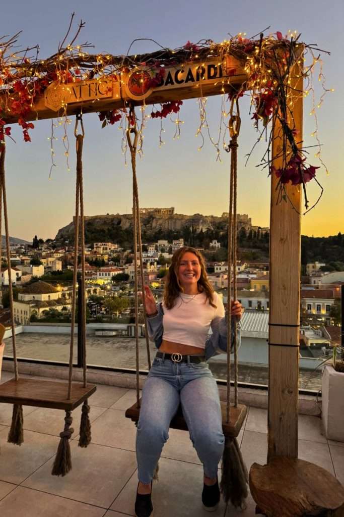 Traveler Liz on a swing with a view that overlooks the Acropolis in Athens during sunset